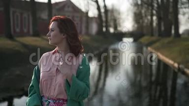 在温暖的春天天气里，穿着绿色时尚夹克的年轻女子在皇宫运河附近放松-红头发